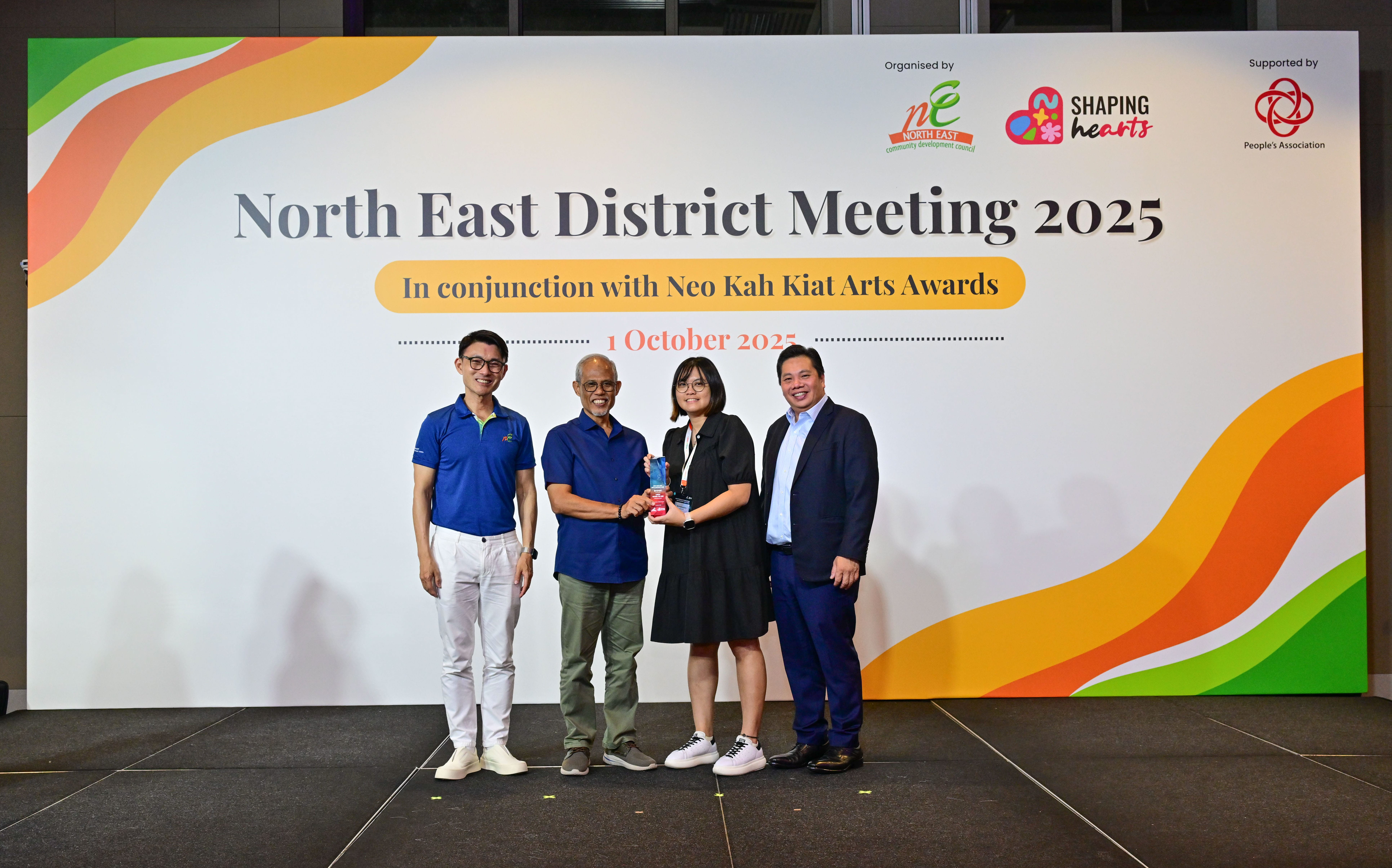 Award recipient standing on stage with the presenter, holding a acrylic plaque and posing for a group photo during the award ceremony, with a backdrop reading ‘North East District Meeting 2025 In conjunction with Neo Kah Kiat Arts Awards'